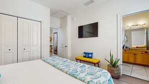 Quarto com uma cama grande, um banco, uma TV e banheiro com espelho e pia.