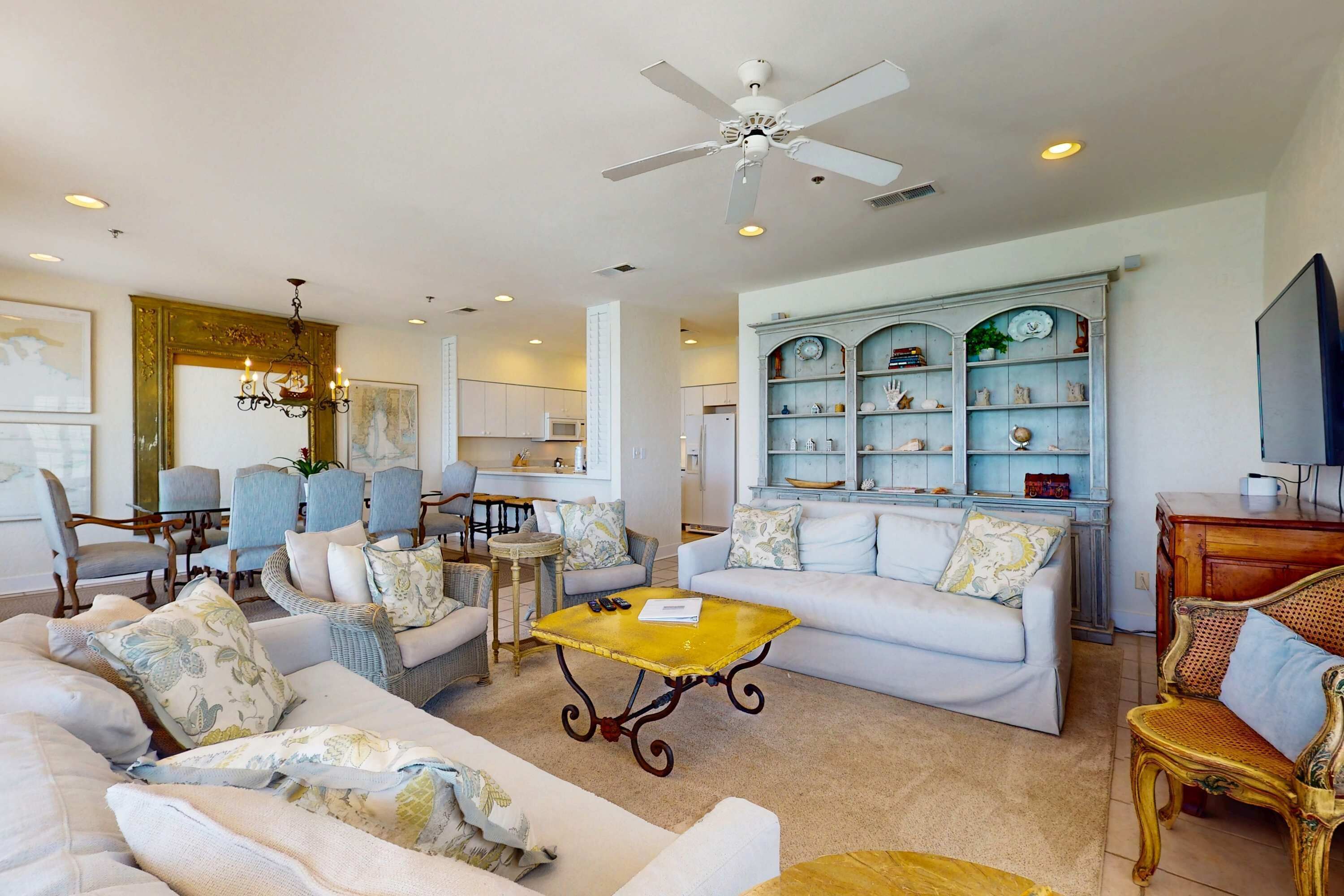 A spacious living room with a white sofa, a yellow coffee table, and a ceiling fan. There is a kitchen in the background with white cabinets and a blue cabinet with decorative items.