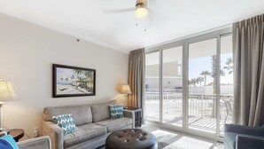 Een moderne woonkamer met een grijze bank, een zwart poef en een schilderij van een strandgezicht.