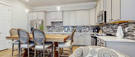 A modern kitchen with white cabinets, a wooden dining table, and a patterned sofa.