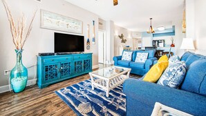 Un soggiorno con un divano blu, un tavolino in vetro, un mobiletti color petrolio e una TV a parete.