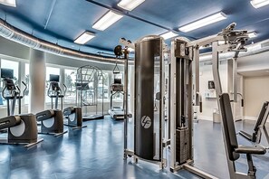 Een sportschool met diverse fitnessapparaten, grote ramen en een blauw plafond.