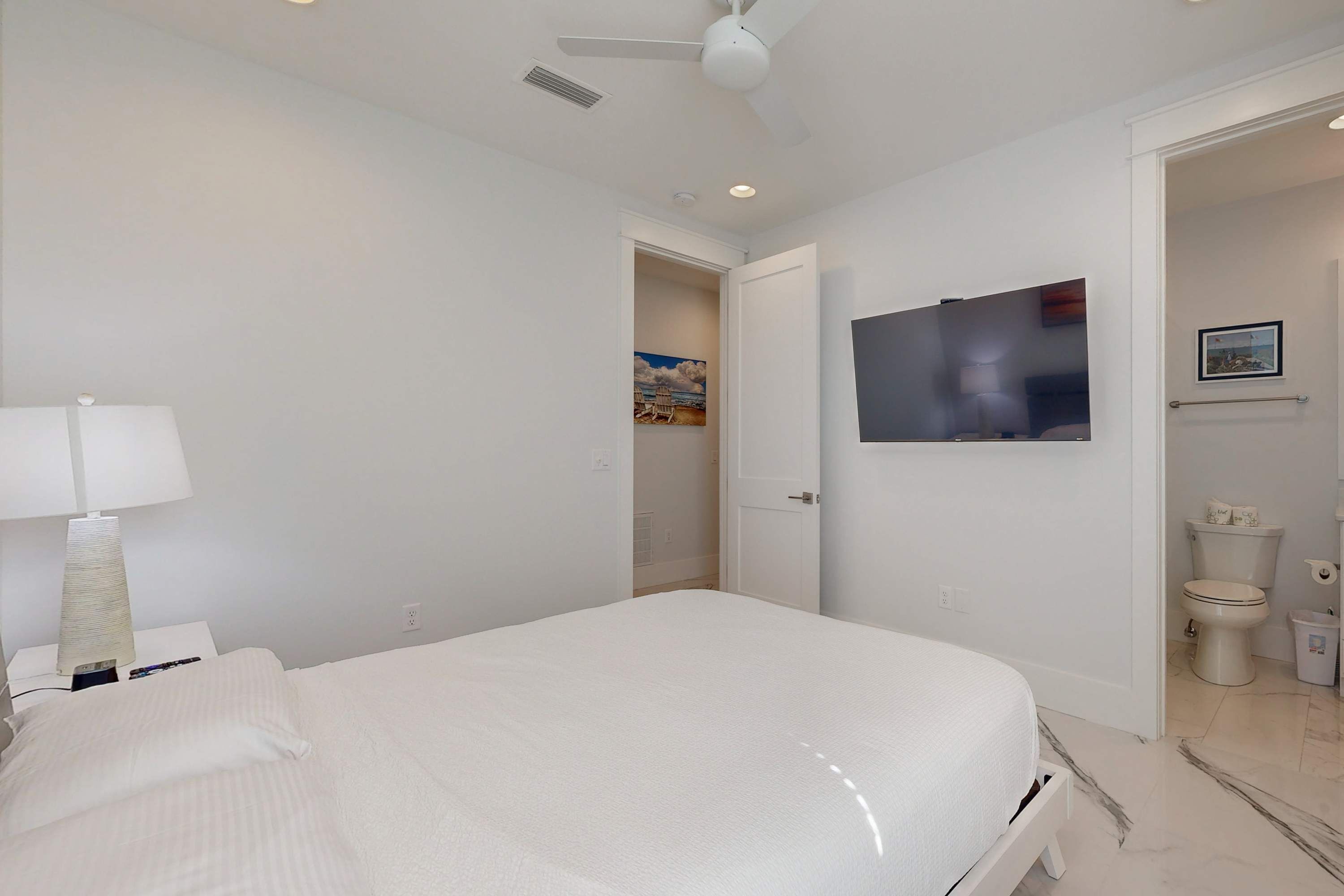 A bedroom with a bed, a nightstand, a lamp, a ceiling fan, a painting, and a view into the bathroom.