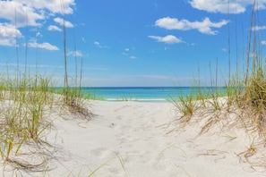 Ein Strand mit weißem Sand und hohem Gras im Vordergrund, der zu einem klaren, blauen Meer unter einem teils bewölkten Himmel führt.
