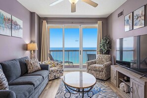 A living room with a sofa, two armchairs, a coffee table, a TV, and a balcony with a view of the ocean.