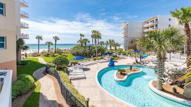 A resort pool area with a curved pool, lounge chairs, and palm trees. - Ground-Floor Condo At Waterscape 490 Feet Of Private Beach (Fort Walton Beach)