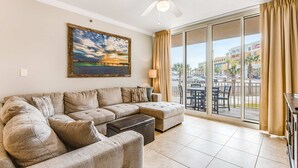 A living room with a sectional sofa, a ceiling fan, a large window with curtains, and a painting on the wall. - Ground-Floor Condo At Waterscape 490 Feet Of Private Beach (Fort Walton Beach)