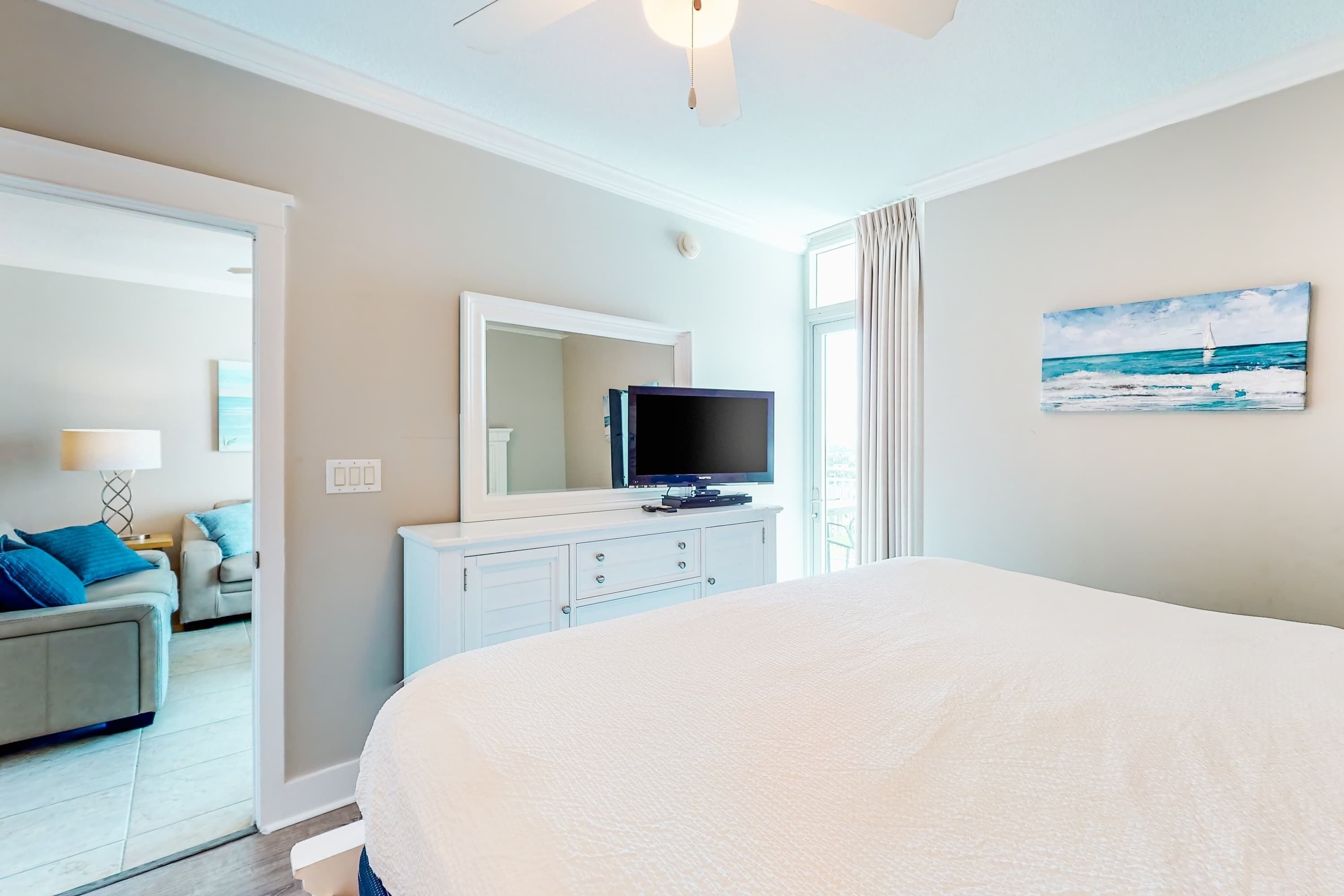 Ein Hotelzimmer mit einem großen Bett, einem Fernseher auf einer Kommode, einem Bild mit Strandmotiv und Blick auf das Meer durch das Fenster.