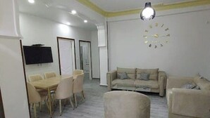 A modern living room with a beige sofa, a round ottoman, a dining table with chairs, a flat-screen TV, and a wall clock. - Whole house: apartment - Béjaïa, Algeria (Béjaïa)
