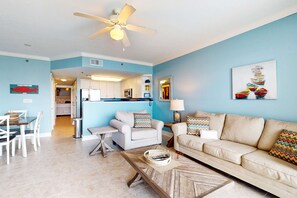 A living room with a beige sofa, a coffee table, and a ceiling fan. There is a kitchen in the background with white cabinets and a microwave. - Waterscape Condo w Shared Amenities, Seasonal Beach Service Included (Fort Walton Beach)