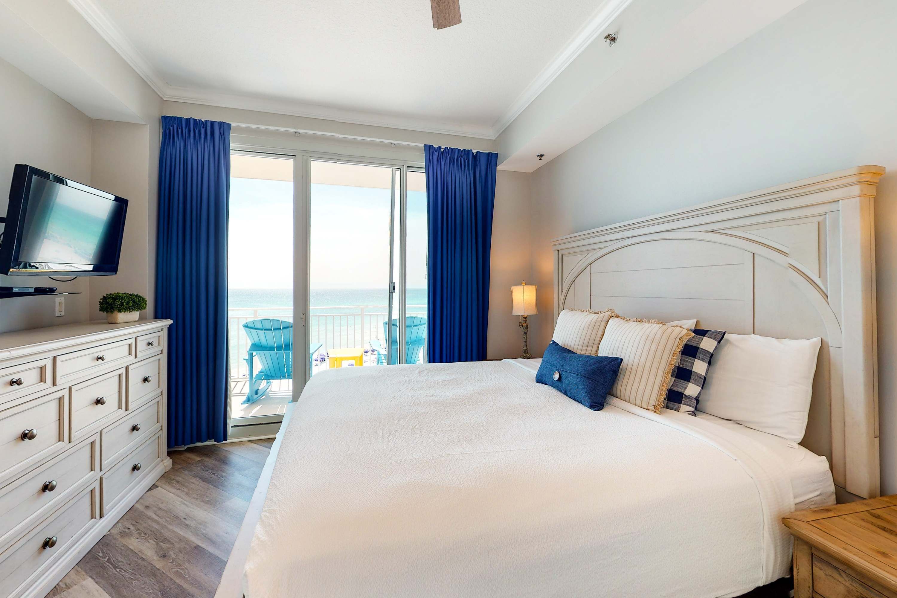 A hotel room with a large bed, a television, a dresser, and a balcony with blue curtains.