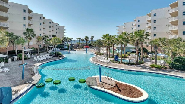 A swimming pool with lounge chairs and palm trees, surrounded by multi-story buildings. - Beach-Chic Condo On Okaloosa Island Steps To Beach Lazy River (Fort Walton Beach)