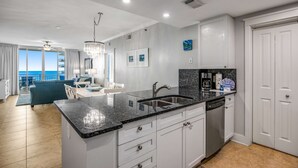 Una cocina moderna con encimera de granito, armarios blancos y lavavajillas. Cuenta con una zona de comedor con mesa y sillas, y vistas al océano a través de amplios ventanales.