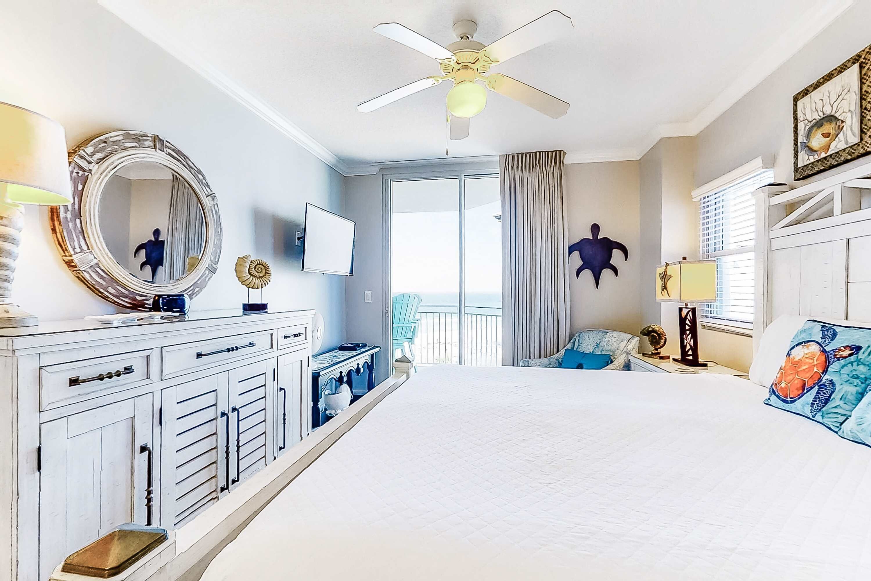 A bedroom with a large bed, a ceiling fan, a dresser, a TV, and a view of the outdoors.