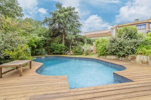 Eine Holzterrasse umgibt einen Swimmingpool mit einer Bank, umgeben von üppiger Vegetation und einem Haus im Hintergrund.