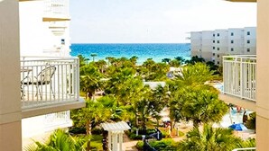 Vista de uma varanda com vista para uma praia, palmeiras e céu azul límpido.