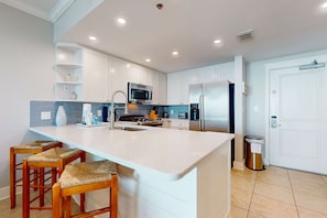 A modern kitchen with white cabinets, a large island with bar stools, stainless steel appliances, and a tiled backsplash. - Inviting, Beach-Chic Condo On Okaloosa Island 490 Feet Of Private Beach (Fort Walton Beach)