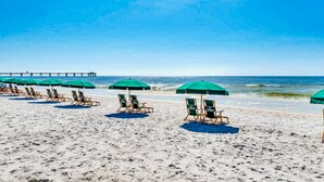 Uma praia com fileiras de espreguiçadeiras e guarda-sóis verdes, um píer ao fundo, e o oceano.