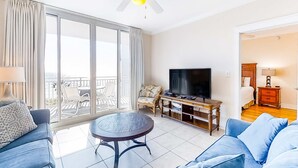 Amplia sala de estar con televisor grande, asientos cómodos, balcón con vistas y ventilador de techo.
