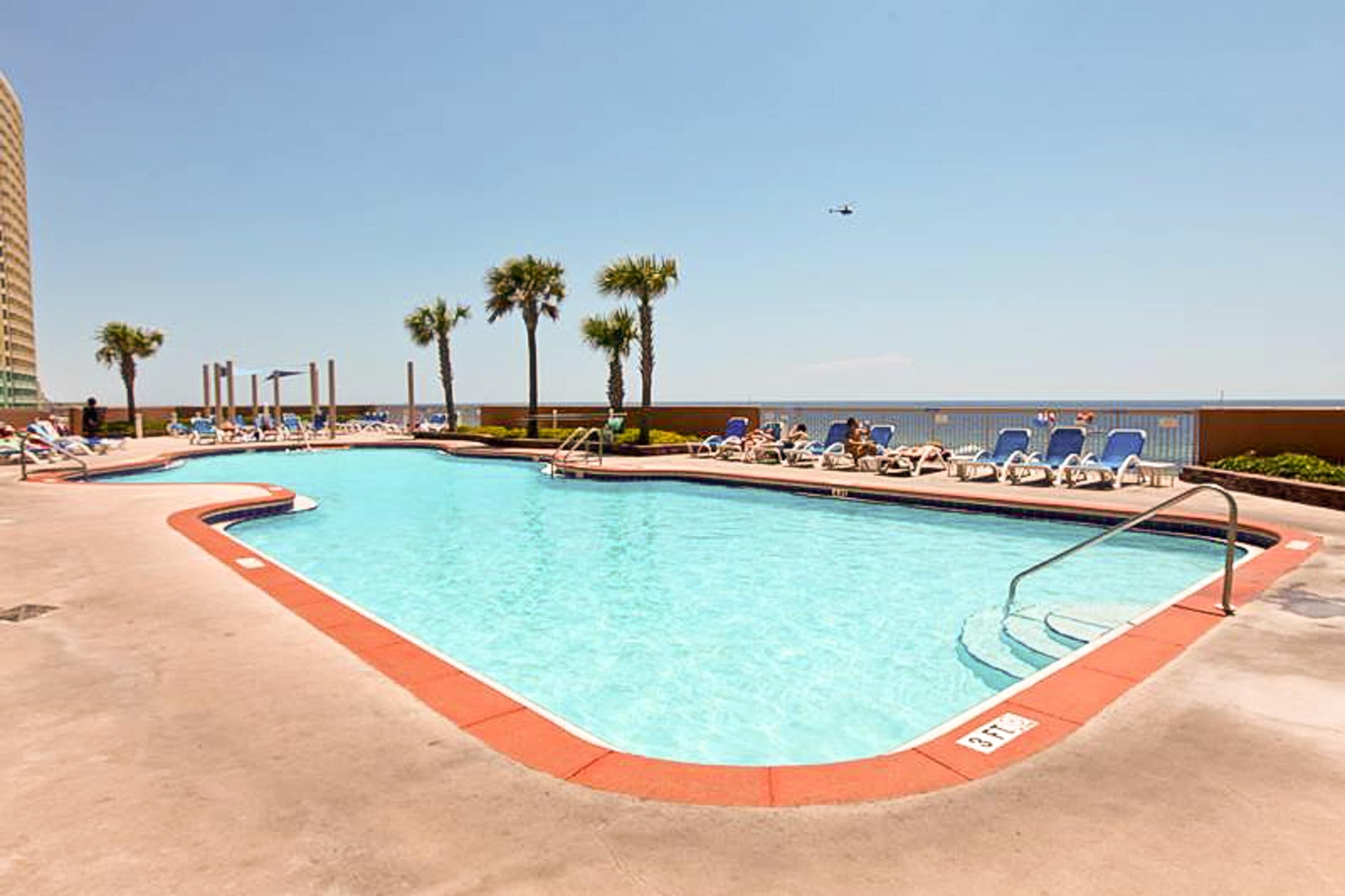 A pool area with lounge chairs and palm trees, a clear sky, and a building in the background.