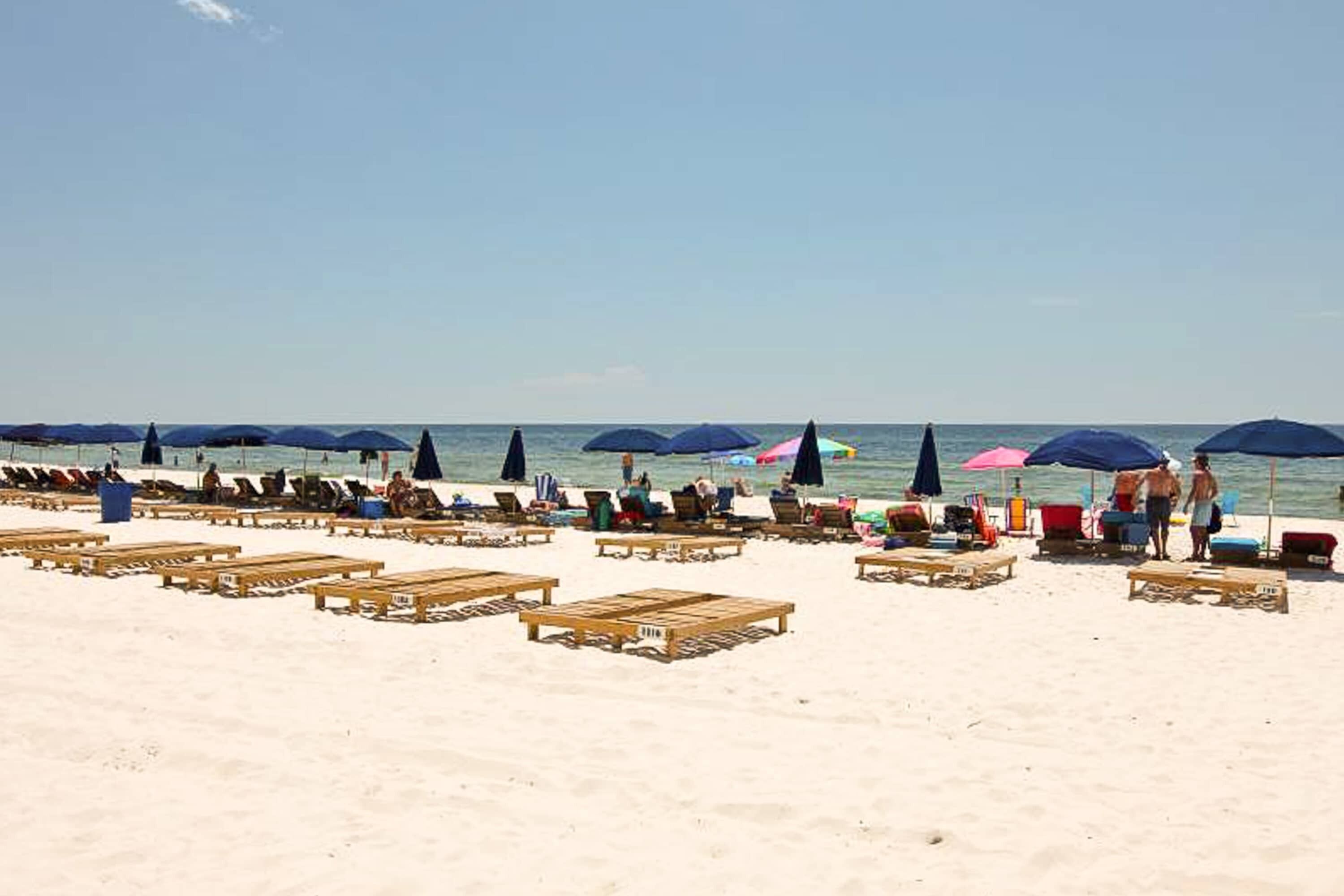 Una playa con hileras de sillas de madera y sombrillas, algunas ocupadas por personas.
