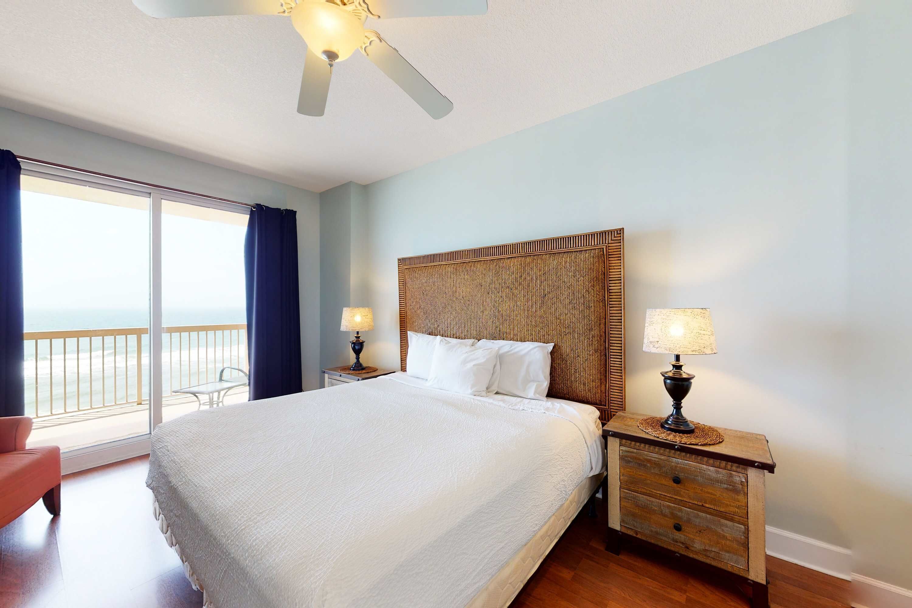 Een hotelkamer met een groot bed, twee nachtkastjes met lampen, een plafondventilator, uitzicht op het strand en een balkon met een stoel.