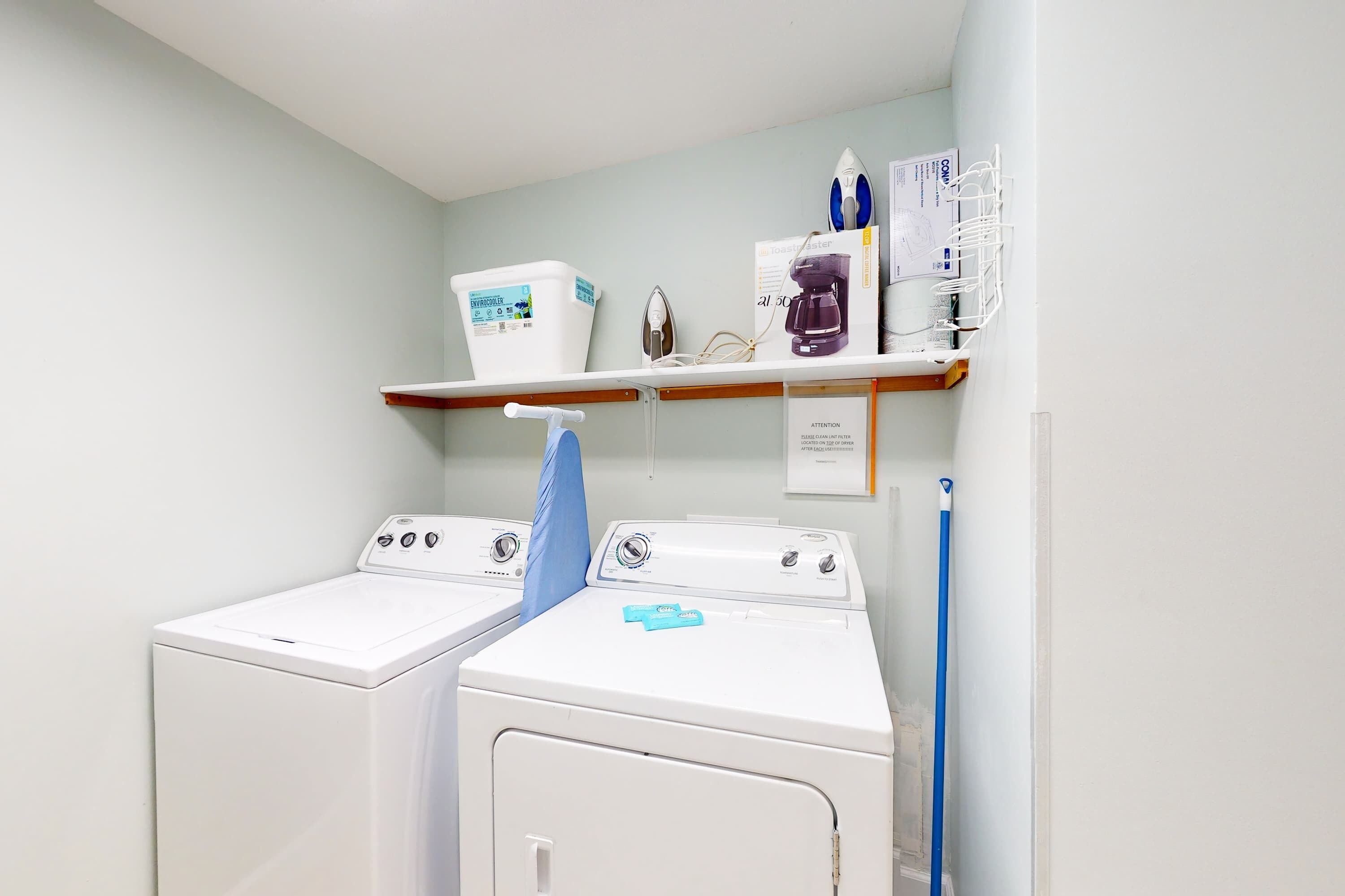 A laundry room with two white front-loading washers and dryers, a blue mop, and a shelf with cleaning supplies.