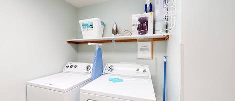 A laundry room with two white front-loading washers and dryers, a blue mop, and a shelf with cleaning supplies.