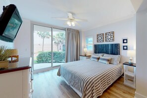 A bedroom with a bed, a ceiling fan, a TV, a window with curtains, and artwork on the wall. - Handicap accessible ground-floor condo on Okaloosa Island Waterfall, Lazy River (Fort Walton Beach)