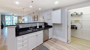 A modern kitchen with white cabinets, black countertops, and a built-in dishwasher. There is a window with curtains, a ceiling fan, and a kitchen island with a sink and a coffee maker. - Handicap accessible ground-floor condo on Okaloosa Island Waterfall, Lazy River (Fort Walton Beach)