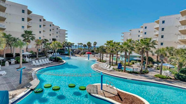 A swimming pool with lounge chairs and palm trees, surrounded by multi-story buildings. - 6th Floor Cozy, 'At Home' Condo At Waterscape Steps To Beach Lazy River (Fort Walton Beach)