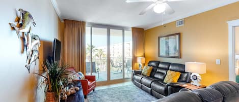 A living room with a leather sofa, a red armchair, a ceiling fan, and a large window with a view of buildings and palm trees.