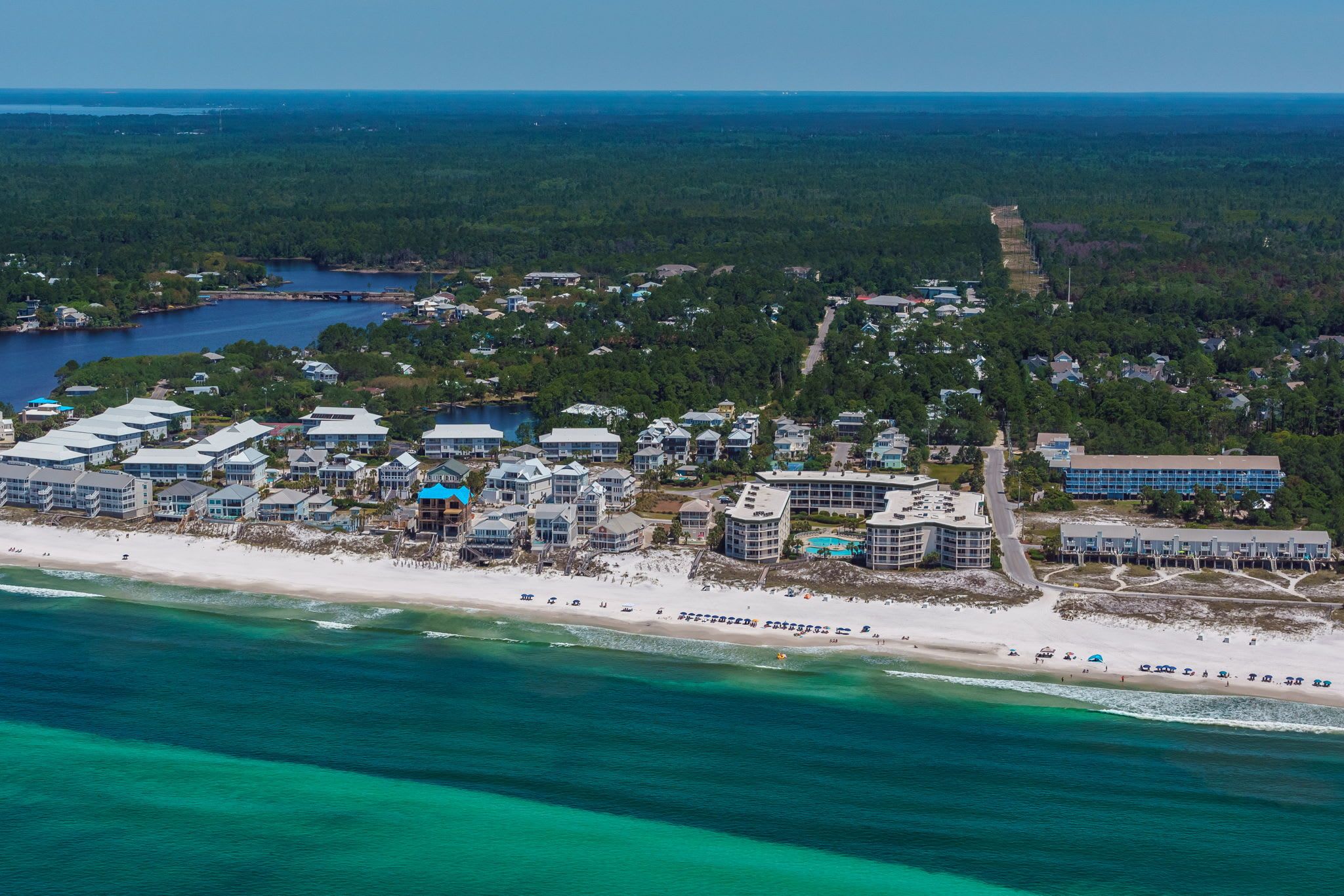 An aerial view of a coastal resort with beachfront properties, a marina, and a dense forest in the background.