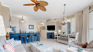 A spacious living room with a sofa, ottoman, and patterned armchairs. A dining area with a bar and chairs is visible in the background. - Beach-view condo with outdoor pool, hot tub, sauna, tennis, BBQ, & beach access (Santa Rosa Beach)