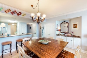 A dining area with a wooden table, bar stools, and a chandelier. - 6th-floor charming condo with amazing views & pools (Perdido Key)