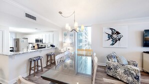 A modern kitchen with white cabinets, a central island, and bar stools. There is a dining area with a glass table and chairs, and a living room with a patterned sofa and a wall-mounted TV. - 4th-floor Gulf-front condo with pools - near entertainment (Perdido Key)