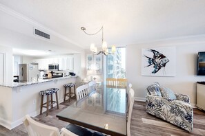 A modern kitchen with white cabinets, a central island, and bar stools. There is a dining area with a glass table and chairs, and a living room with a patterned sofa and a wall-mounted TV. - 4th-floor Gulf-front condo with pools - near entertainment (Perdido Key)