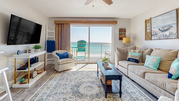 A living room with a TV, a sofa, a coffee table, and a view of the ocean through a sliding door. - Oceanfront condo with pool, hot tub, central AC, washer&dryer, & fitness room (Fort Walton Beach)