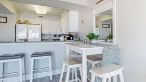 A modern kitchen with white cabinetry, a central island with stools, and a refrigerator with a 'hello' sign. - Oceanfront condo with pool, hot tub, central AC, washer&dryer, & fitness room (Fort Walton Beach)