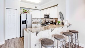A modern kitchen with white cabinets, a granite countertop, a stainless steel refrigerator, a microwave, a stove, and three bar stools. - Gulf-Front Condo w/ 14th-Floor Views, Beach Service & Shared Pools/Hot Tubs (Panama City Beach)