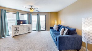 A living room with a blue sofa, a white cabinet, a ceiling fan, and a large window with curtains. - Cozy condo in paradise with pool, sauna & private dock - snowbird-friendly (Panama City Beach)