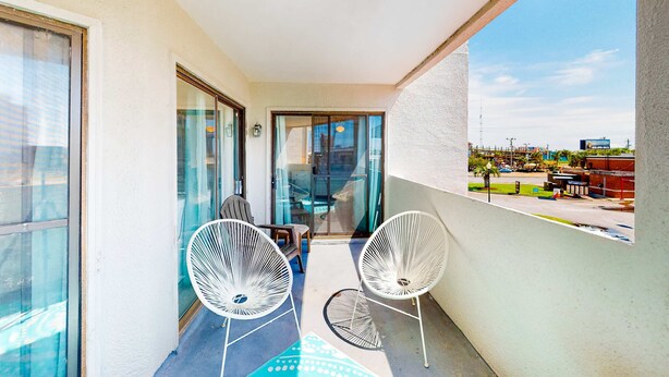 A balcony with two white wire-framed chairs, a glass door leading inside, and a view of a commercial area with buildings and a parking lot. - Cozy condo in paradise with pool, sauna & private dock - snowbird-friendly (Panama City Beach)