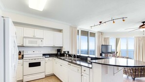 A modern kitchen with white cabinetry, a built-in oven, microwave, and dishwasher. There is a large island with a black countertop and a view of the balcony. - 16th-floor waterfront condo with fast WiFi, Gulf view, pools, hot tubs, & gym (Panama City Beach)