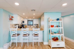 A modern kitchen with white cabinets, a wooden countertop, and a white island with bar stools. There is a white shelving unit with decorative items on the right side. - Charming & elegant Gulf-front condo w/shared pools, hot tub, gym (Panama City Beach)