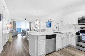 A modern kitchen with white cabinetry, a dishwasher, an oven, and a dining area with a view. - Beachfront penthouse with 2 free beach chairs, WiFi, amazing views, pool & hot tub (Panama City Beach)