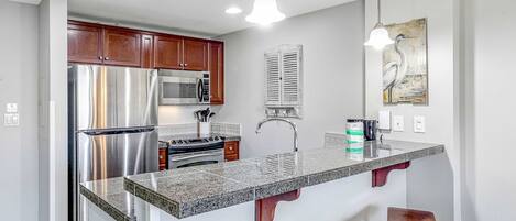 A modern kitchen with stainless steel appliances, a granite countertop, wooden cabinets, and pendant lighting.