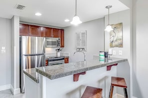 A modern kitchen with stainless steel appliances, a granite countertop, wooden cabinets, and pendant lighting. - Vibrant seventh-floor condo with pool, hot tub, sauna, private W/D & fast WiFi (Miramar Beach)