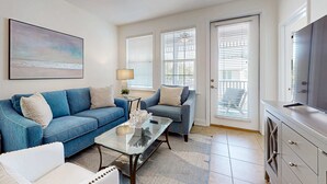 A living room with a blue sofa, a white armchair, a glass coffee table, and a TV on a cabinet. There are two windows with blinds, a ceiling fan, and a painting on the wall. - Blue Mountain Beach – charming 2nd floor condo with pool view (Santa Rosa Beach)