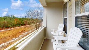Een balkon met witte Adirondack-stoelen, een kleine tafel en uitzicht op een weg en bomen.