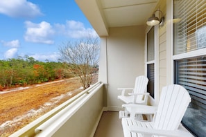 A balcony with white Adirondack chairs, a small table, and a view of a road and trees. - 2nd-floor condo near beach with balcony, outdoor pool, streaming, & washer/dryer (Santa Rosa Beach)